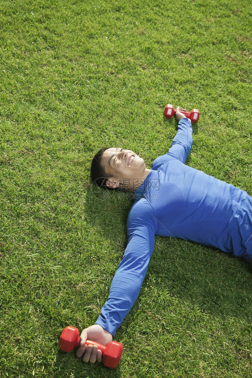 年轻的运动健壮的年轻人躺在公园里 在草地上与哑铃一起躺着图片
