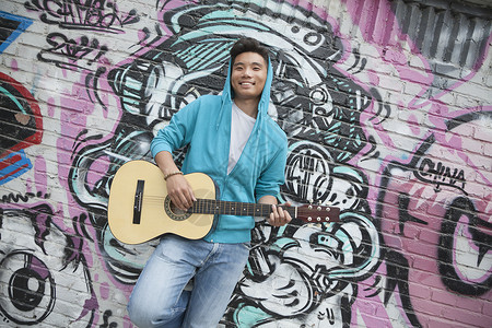 年轻 笑着的街头音乐家靠在墙上 涂鸦画画和弹吉他服装乐趣享受幸福音乐原声摄影娱乐露齿城市生活背景图片