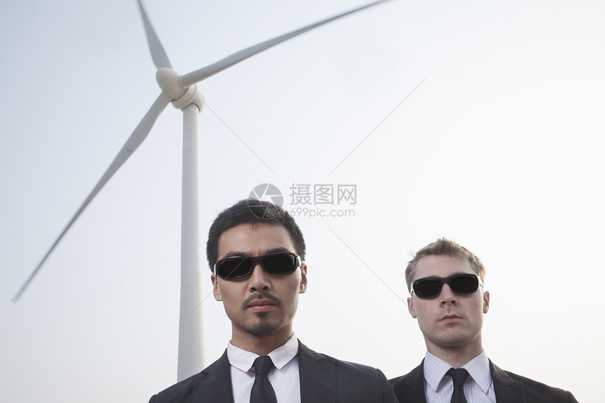 两名身戴太阳镜的严肃青年商务人士在风力涡轮机旁站立的肖像图片