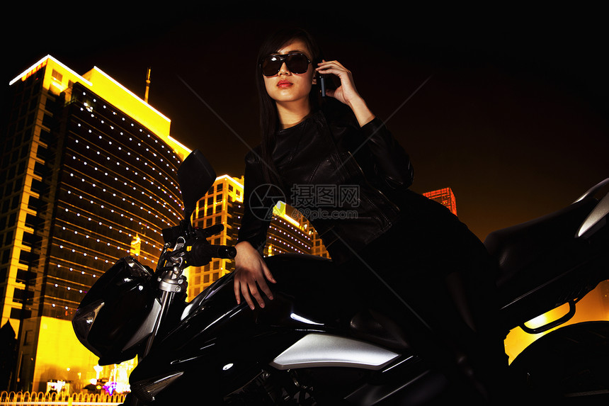 美丽的年轻女子 带着太阳眼镜 在电话上说话 晚上靠着摩托车在夜里倾斜女性沟通方式太阳镜体力运输建筑手机活动冒险图片