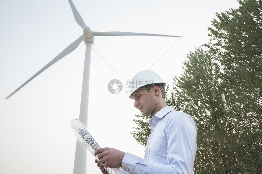 年轻男工程师身穿硬马帽 看着风力涡轮机前的蓝图图片