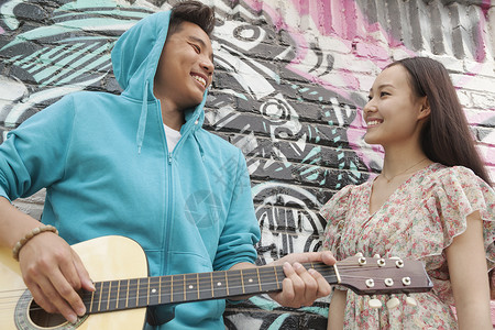 年轻的微笑街头音乐家靠在涂鸦画的墙上 弹着吉他 和一个穿着裙子的年轻女人调情背景图片