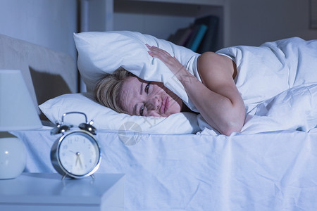 妇女用枕头顶住耳朵 作为闹钟铃响女性女士卧室床单桌子家庭生活软垫睡眠苏醒毯子背景图片