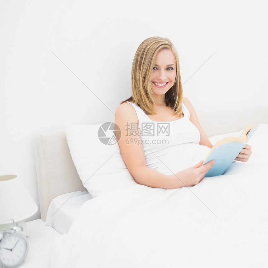 幸福女人在床上看书的肖像枕头闹钟文学微笑头发床头柜毯子女士金发女郎棉被图片