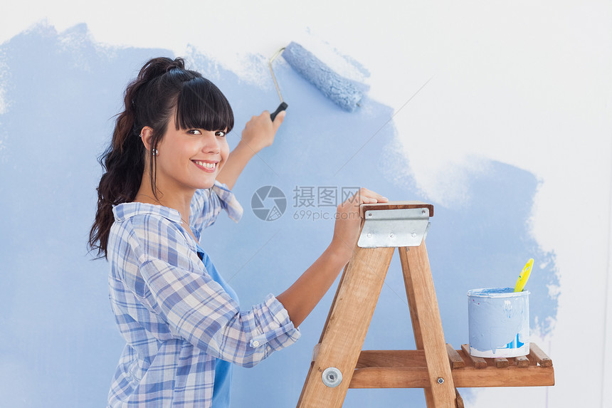妇女用涂漆滚滚来油漆墙壁 对着照相机微笑图片