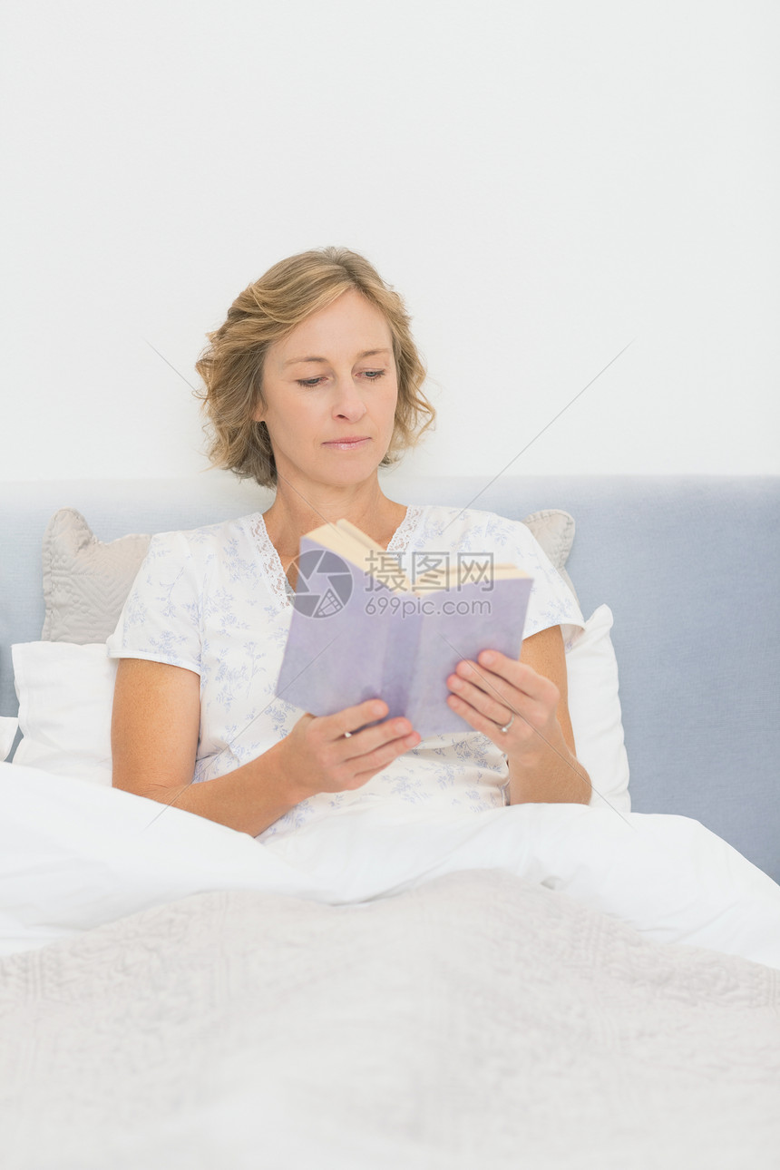 坐在床上读书的金发女人被子房子短发枕头金发女郎住所坐垫微笑家庭卧室图片