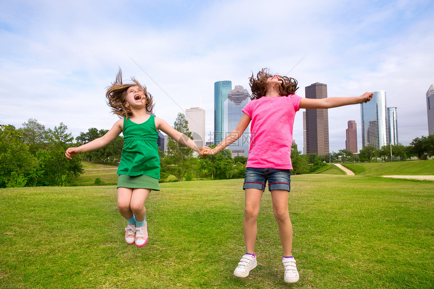 两个女孩在城市天际上 握着手 快乐地跳过两个朋友女孩们草地公园孩子们金发姐妹跳跃地标朋友们高楼图片