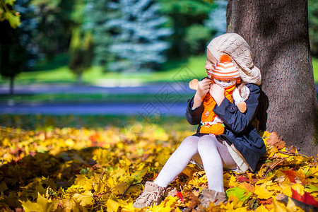 拿着树叶孩子美丽的时装女郎 在阳光明媚的秋天坐在一棵有洋娃娃的山坡树下树木晴天乐趣树叶森林女孩橙子天气童年玩具背景