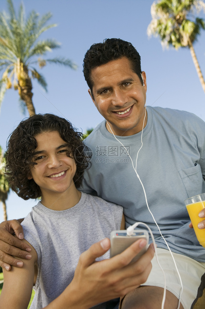 男孩(13-15)持便携音乐播放器的父亲听耳机 并拿着一杯果汁前观景肖像图片