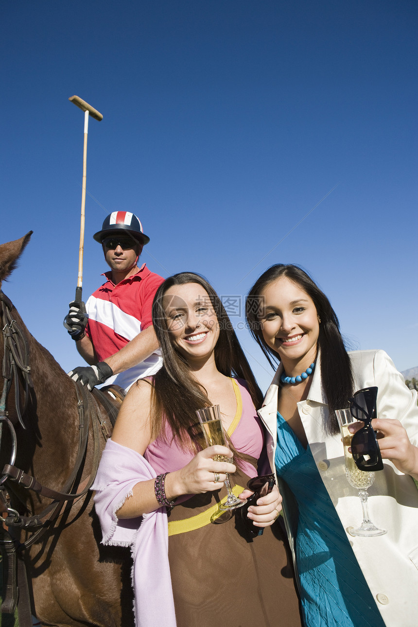 快乐的女朋友和马球手一起喝香槟的肖像 在背景中坐在马背上图片