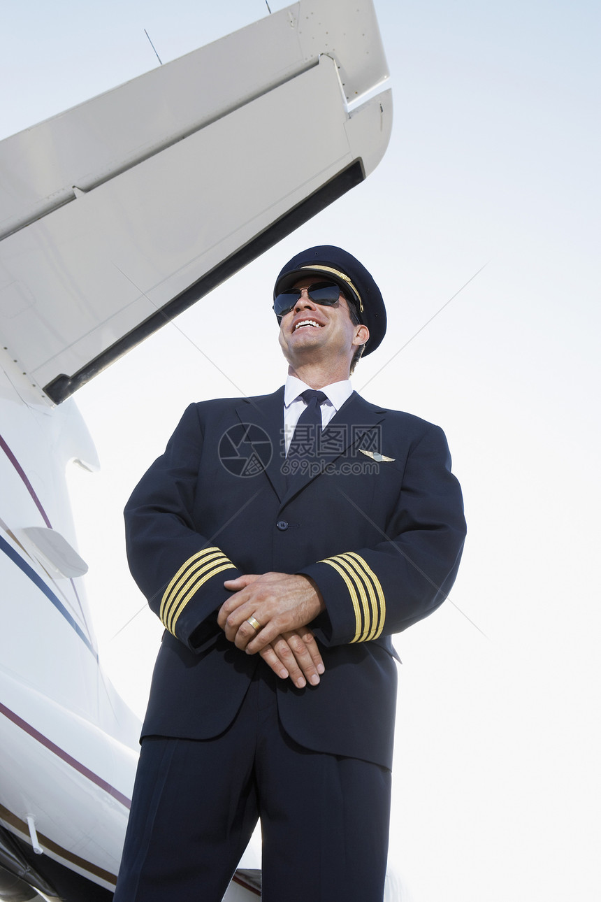 一个穿着制服的笑笑驾驶员 站在手拍着飞机旁边的低角度视角图片