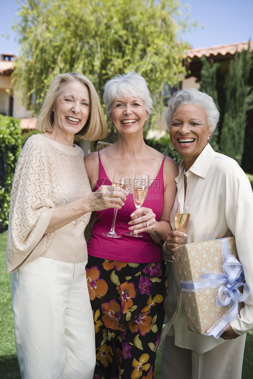 三名多族裔妇女与在草坪的香槟杯一起站在一起的肖像图片