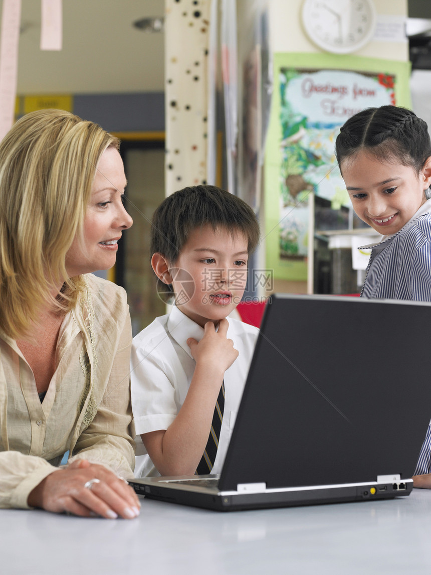 教师用笔记本电脑坐在教室桌上的书桌旁与儿童一起坐着的教师图片
