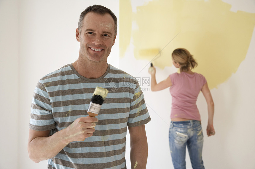 微笑的男人拿着油漆刷子 而妇女则在背景中用滚滚墙壁作画图片