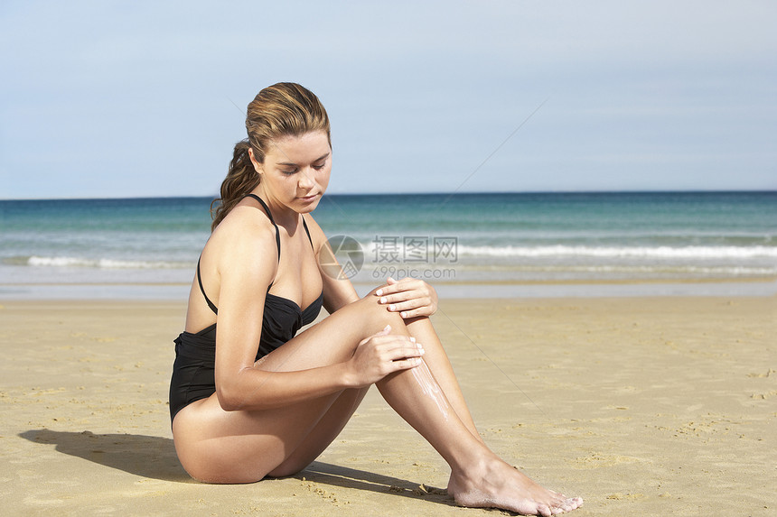 美丽的年轻女子坐在沙沙滩上 在腿上涂防晒霜图片