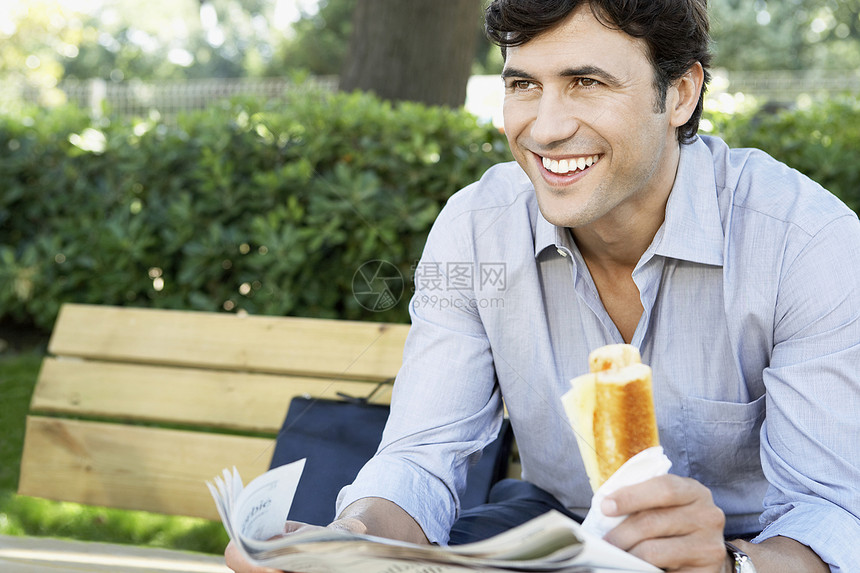 快乐的年轻商务人士 带着三明治和报纸在公园坐在长凳上时看不着边看图片