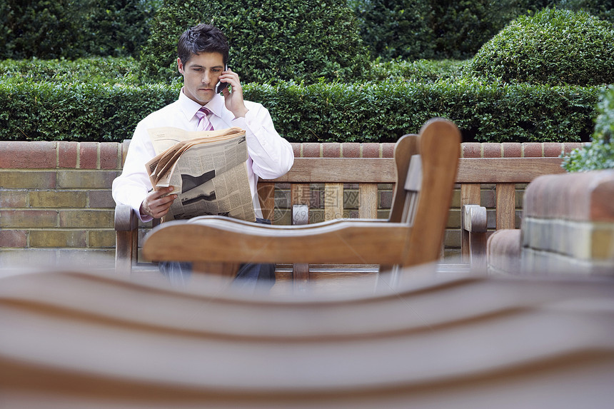 青年商务人士在公园长凳上阅读报纸时使用移动电话;和图片