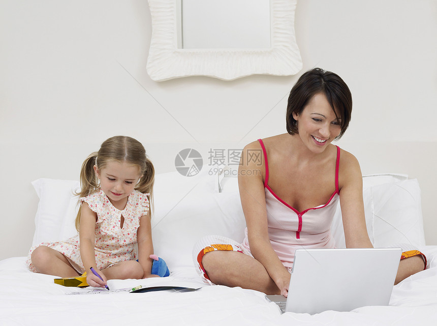 母亲在床上画画时使用女儿的笔记本电脑计算享受技术微笑场景创造力闲暇女孩绘画网络图片