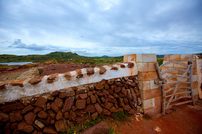 在Menorca带栅栏门的红披萨红色石墙草原场地牧草季节阳光石工岩石植物草地荒野图片