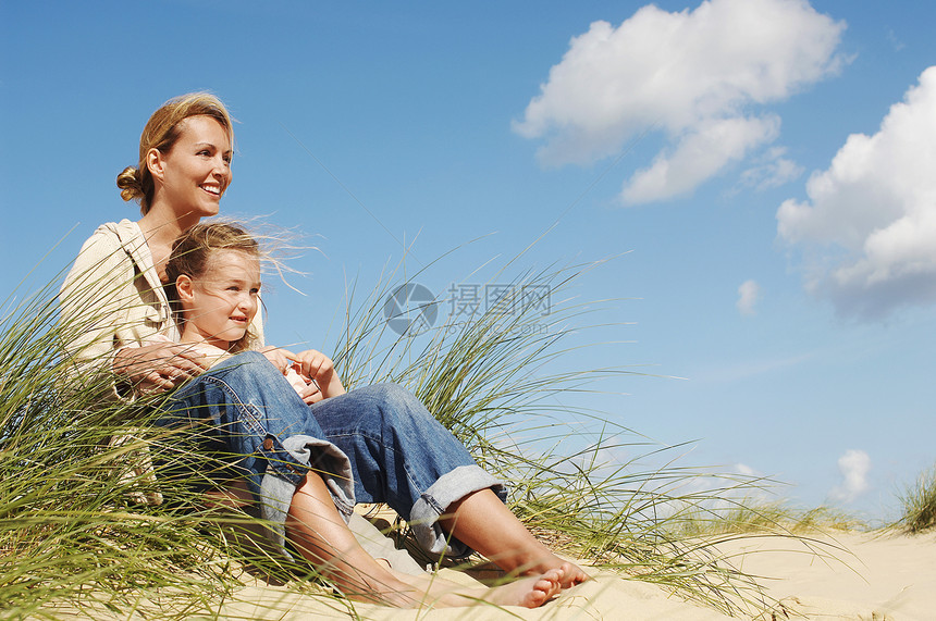 快乐的母亲和小女儿坐在沙滩上的沙地上自由家长孕产天空女孩旅行空闲幸福成人家庭图片