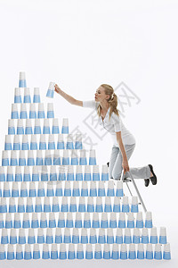飘摇坐在梯子上的年轻女子的侧面景观 将塑料杯叠成金字塔和白色背景背景