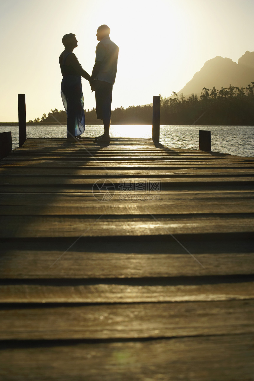 日落时在湖边码头边缘站立的快乐老年情侣的侧景图片