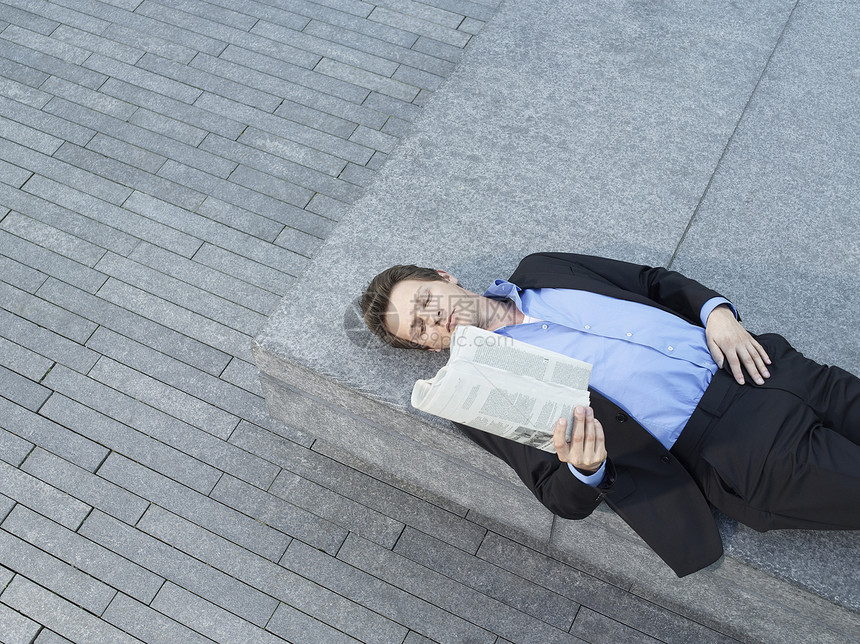躺在人行道上墙壁上的青年商务人士阅读报纸高视率图片