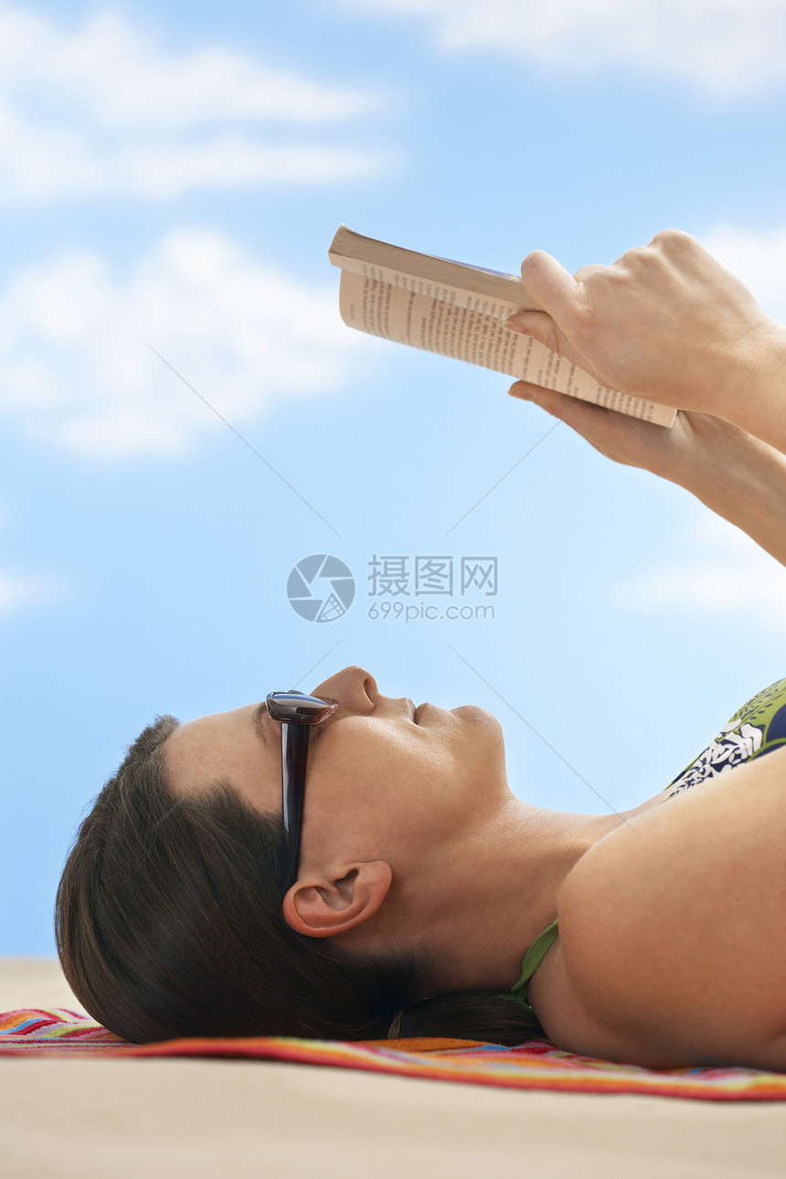 在海滩上读书时 身戴墨镜的年轻女子的侧面观图片
