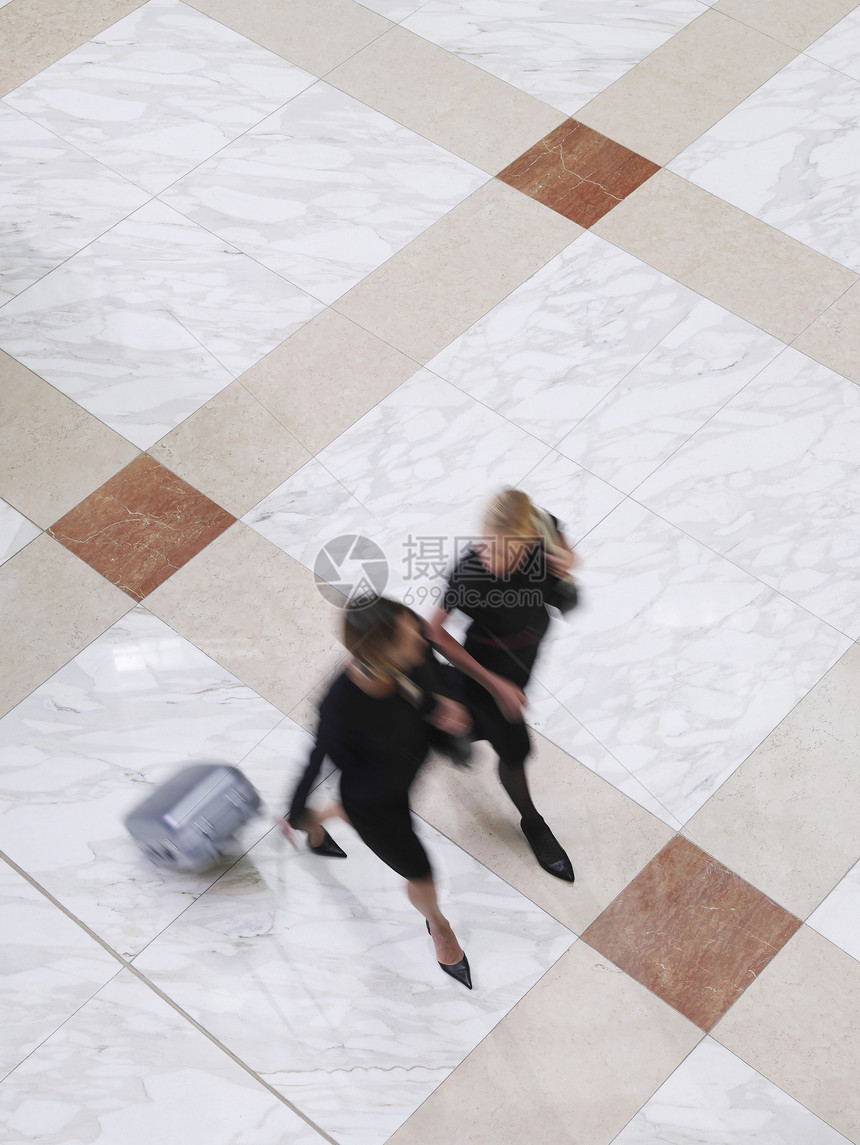 两名模糊的女商务人士在瓦高地板上带着手提箱行走的高视线图片