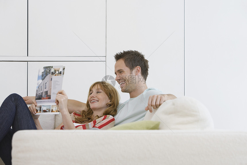 一对快乐和放松的年轻夫妇在沙发上阅读小册子的侧面观点团结成人微笑展柜目录销售夫妻住房房子宣传册图片