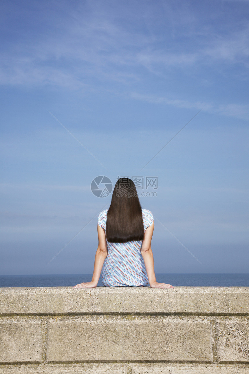 年轻女子坐在海滩的石壁上 与蓝天对立图片