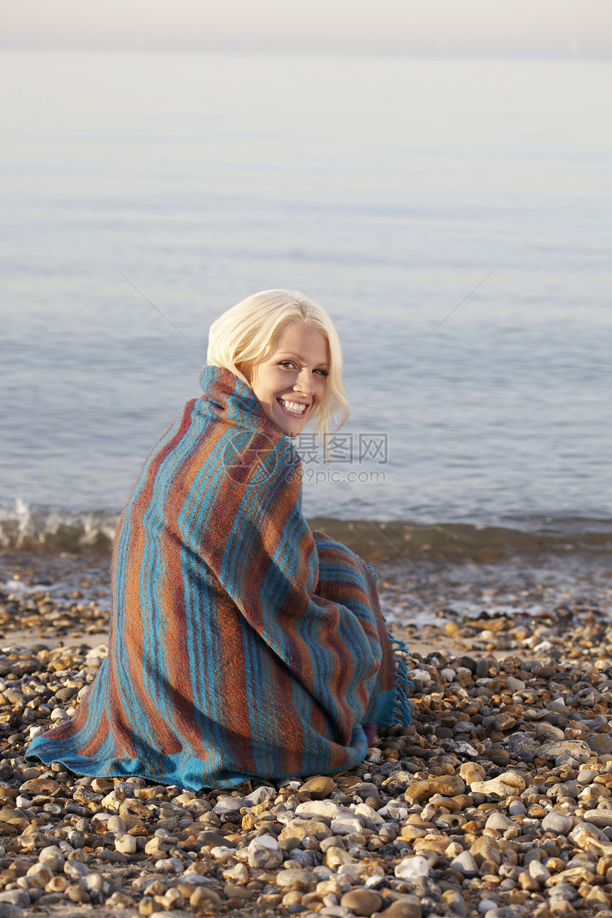 美丽的年轻女人的肖像 被裹在毯子里坐在沙滩上图片