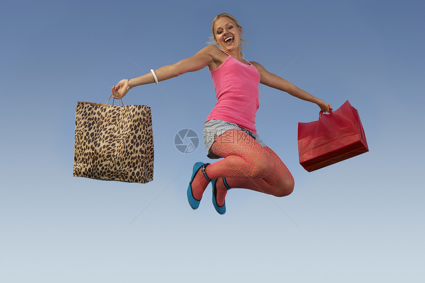 一名兴奋的年轻女子在蓝天上拿着购物袋跳跃的肖像图片