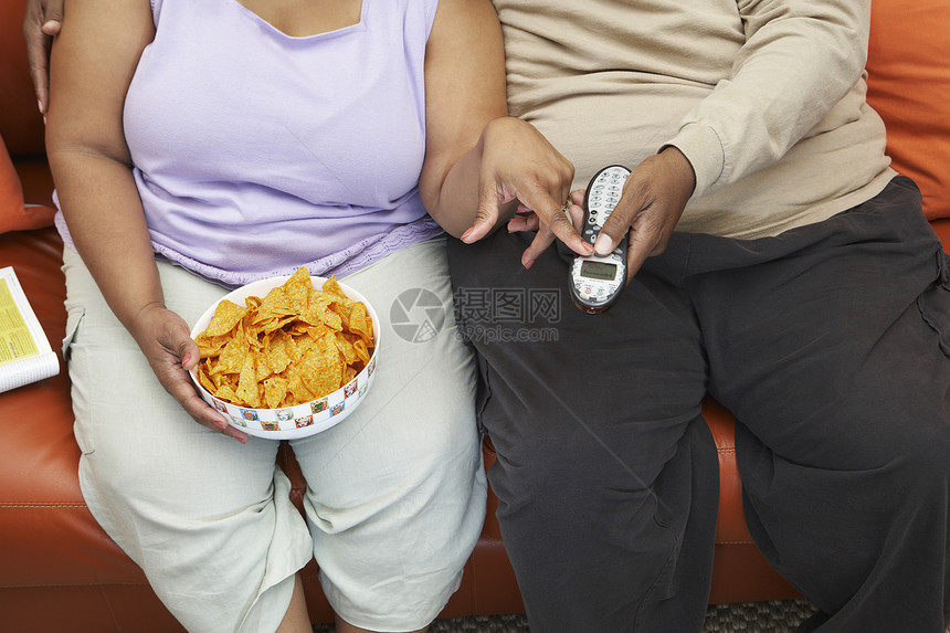 坐在沙发上的一对肥胖夫妇的中段 他们拿着玉米片和遥控器图片