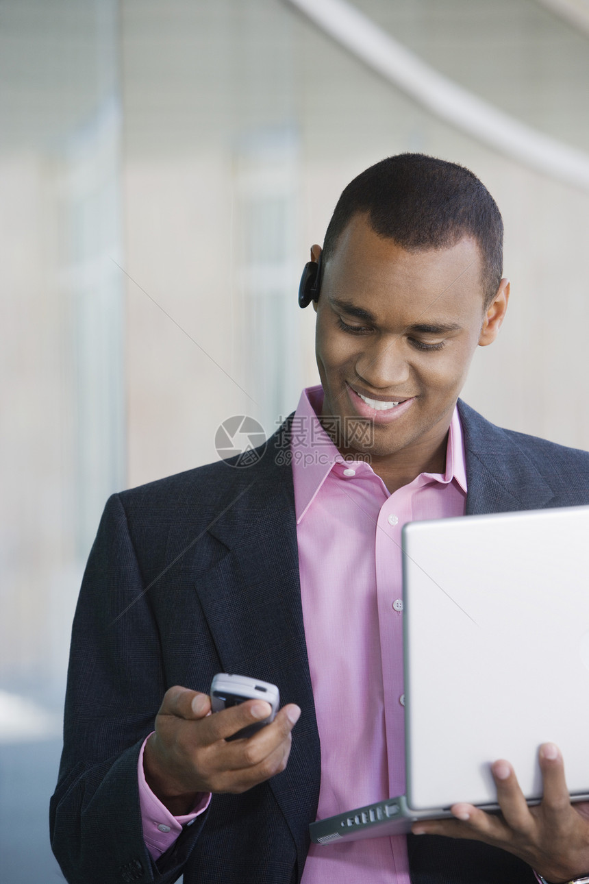 一名非裔美国商务人士持有手提笔记本电脑时在移动电话上阅读短信的讯息图片