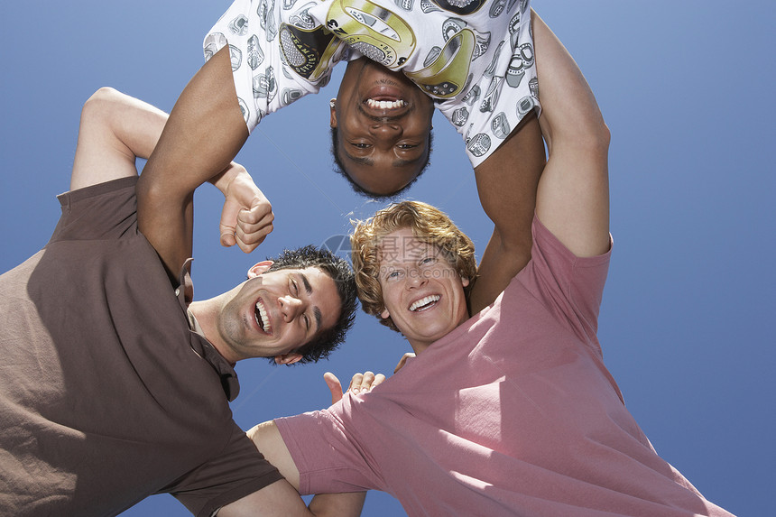 三个快乐的多种族男性朋友在蓝天上交织在一起的低角度视角图片