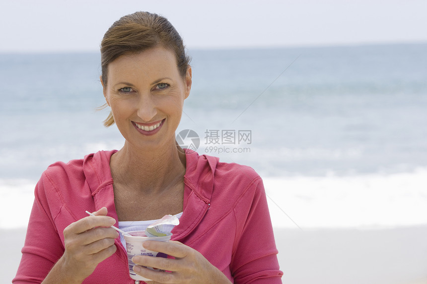 女人在海滩上吃酸奶快乐头发帽衫享受微笑海洋健康饮食粉色外表上衣图片