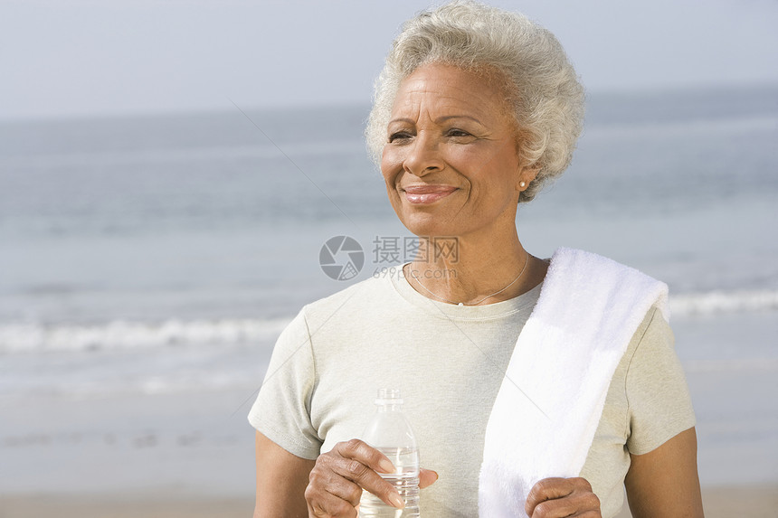 在海滩上 高级妇女用饮用水和毛巾站在她肩上的沙滩上图片