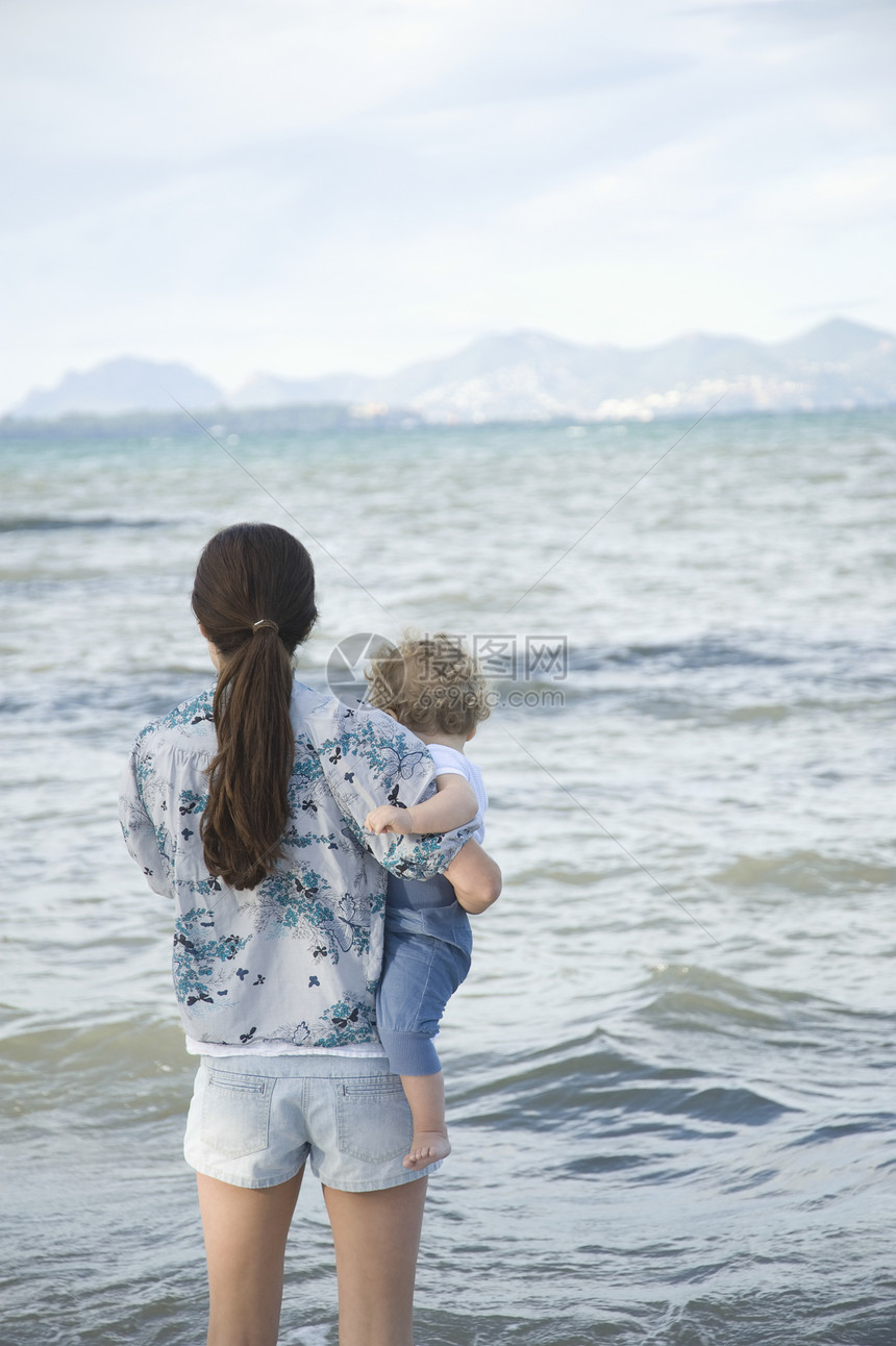 一位女士拿着一个幼儿手站在海面前 有着远处的山峰背景图片