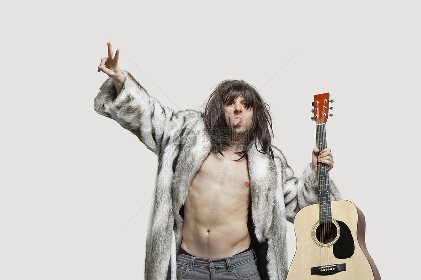 穿毛皮大衣的年轻人拿着吉他的吉他 在灰色背景上巡视图片