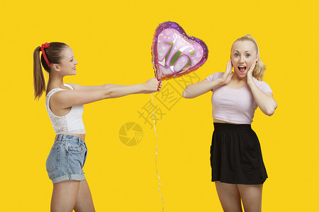 快乐的年轻女子给一个生日气球 给那些站在黄色背景上的惊人女人女性长度派对微笑嘴巴惊喜成人服装影棚两个人背景图片