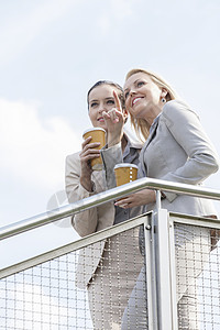 拥有可支配咖啡杯的年轻女商务人士与同事站在一起 靠着天搭扶栏木桩背景图片