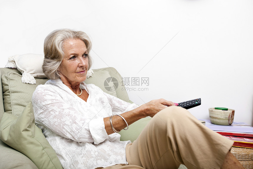高级妇女在家里用遥控器 同时在扶手椅上放松图片