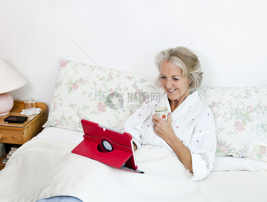 快乐的老年妇女在家里用数字平板电脑 同时在床上喝咖啡图片