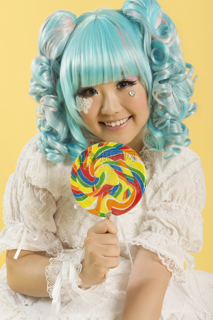 微笑的年轻女子穿成一个洋娃娃 把棒棒棒糖放在黄色背景之上的肖像图片