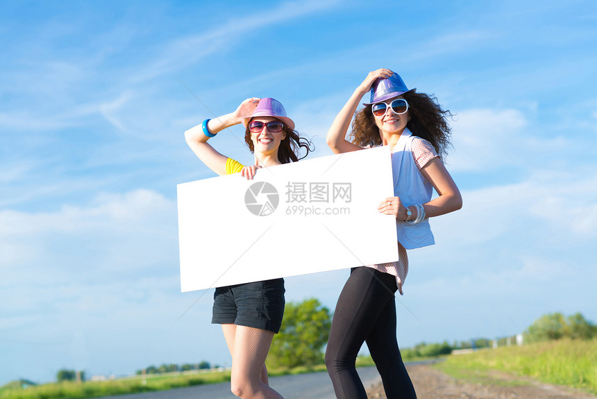 两名年轻女性站在空白的横幅上站着便车航程旅游女士女孩假期木板纸板天空成人图片