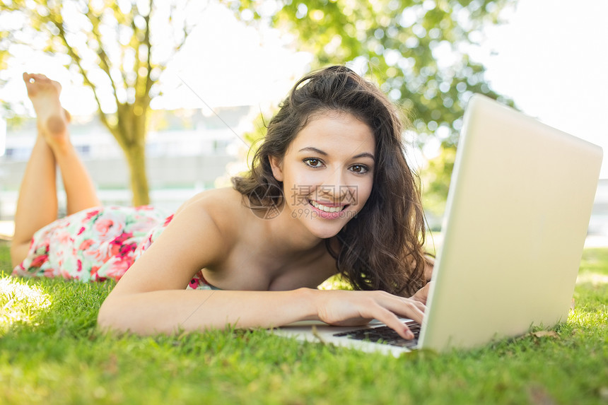 利用她的笔记本电脑躺在草地上的时髦的笑脸黑发美女图片