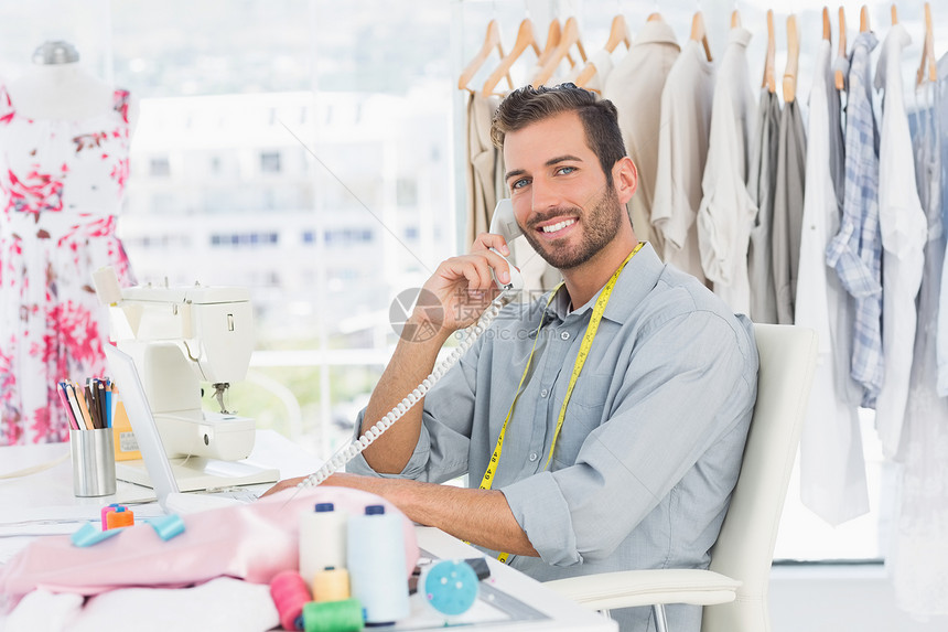 年轻男性时装设计师使用电话的肖像工艺造型师桌子工作固定电话材料呼唤裁缝商业男人图片