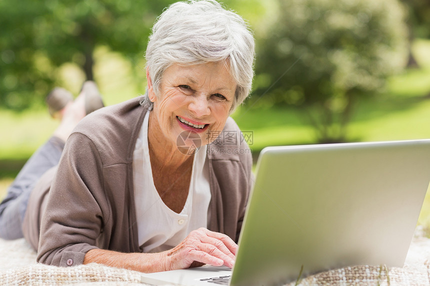 在公园使用笔记本电脑的老年微笑妇女技术说谎快乐女性女士退休头发人员地毯成人图片