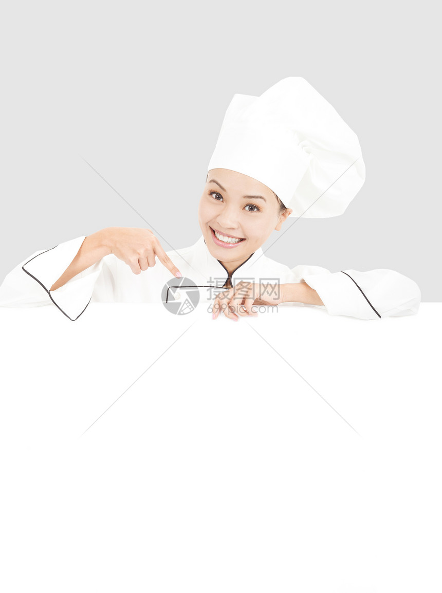 微笑的年轻女厨师用空白板指着工作室女性女士帽子成人商业食物烹饪女孩围裙图片